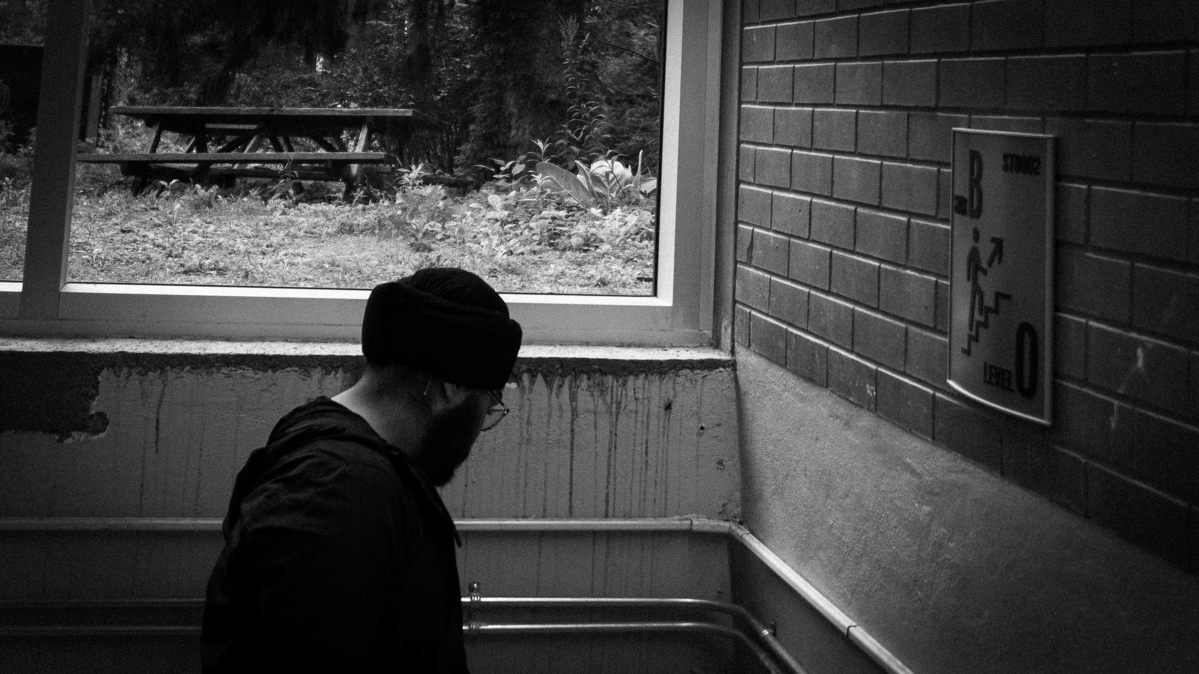 Suraj, a turbaned man, with a short beard, standing looking down in a stairwell, surrounded with concrete walls. There appears to be a window just above his head, filled with grass and a bench.
